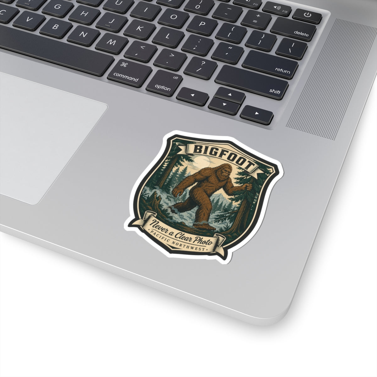 Small Bigfoot hiking badge sticker on laptop lid, brown Sasquatch forest shield artwork