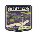 Die-cut sticker showing gray alien walking on a desert road with a hovering UFO.