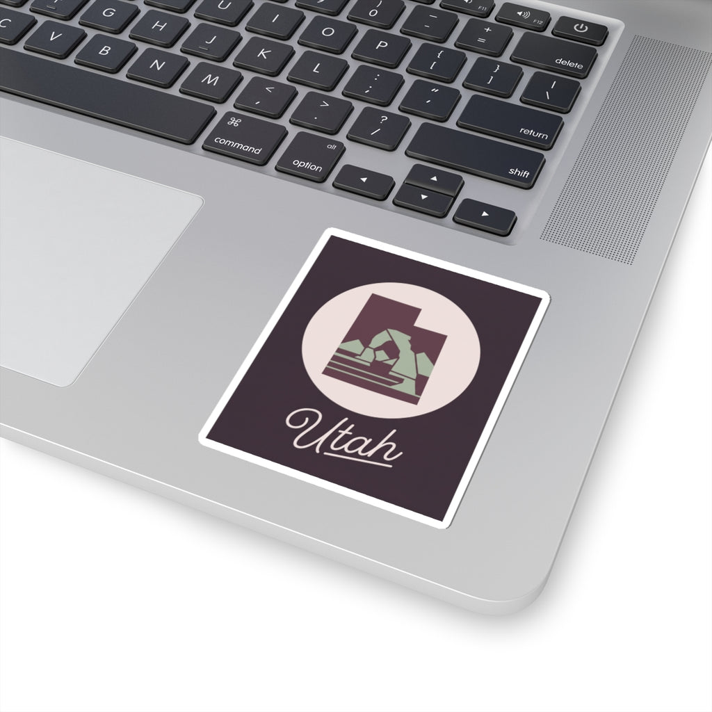 Utah state kiss-cut decal sticker on a laptop, dark purple square with circular mountain and “Utah”.