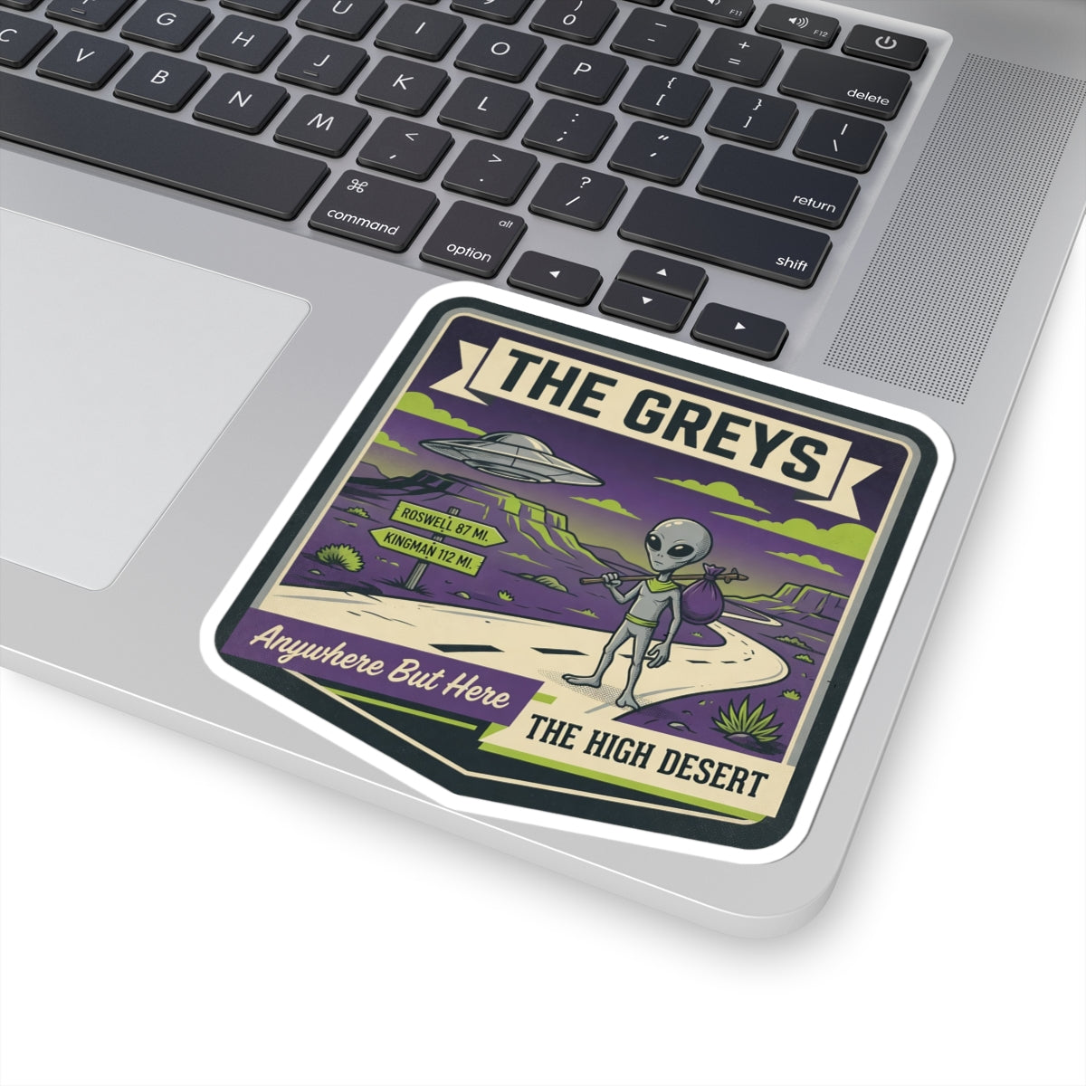 Die-cut laptop sticker with "THE GREYS" title, gray alien, UFO, and desert road.