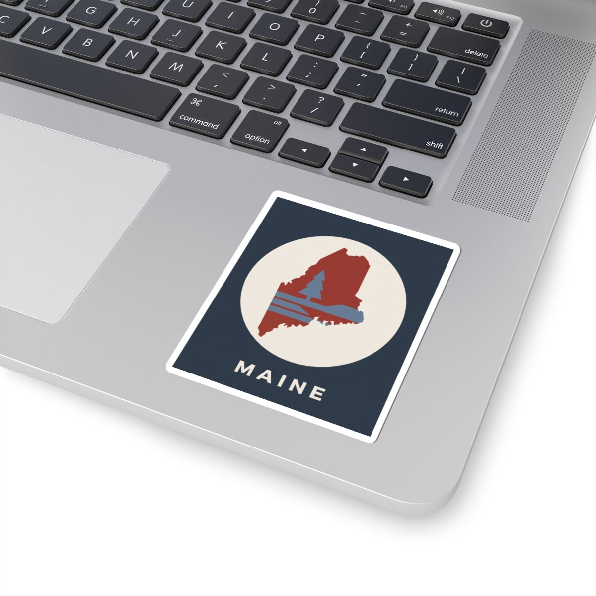 Maine state silhouette sticker on laptop: navy square with red map graphic and “MAINE”