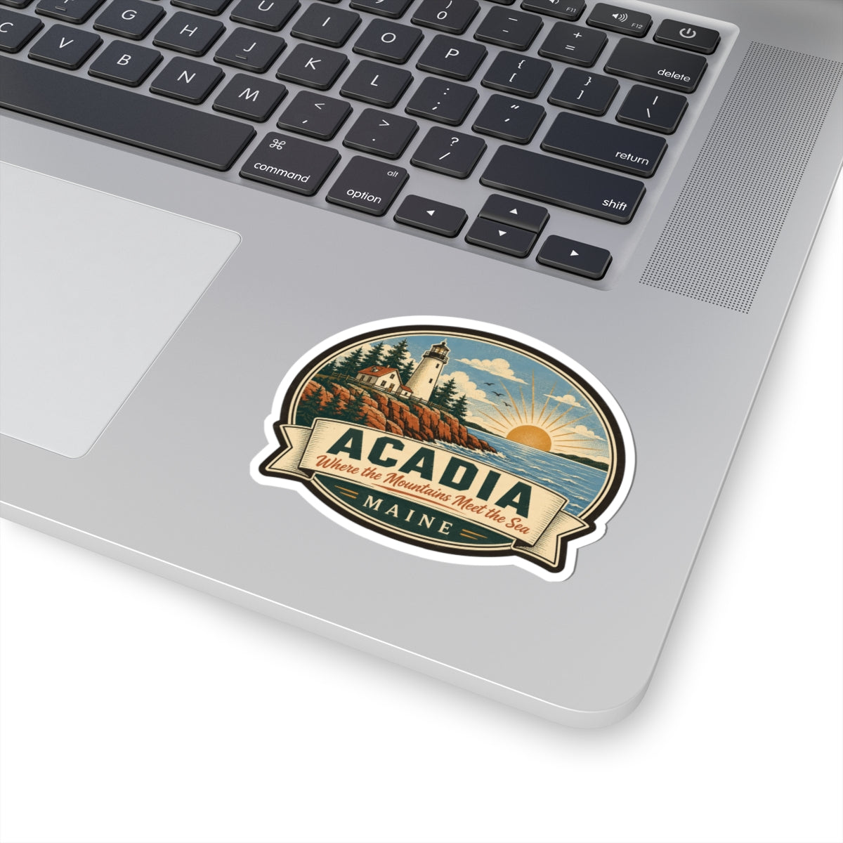 Round Acadia Maine sticker on silver laptop: lighthouse, coastline, sunrise, and 'MAINE' text.