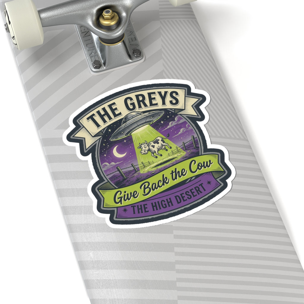 Die-cut sticker on a striped notebook page: UFO and cow, reads 'THE GREYS' and 'THE HIGH DESERT'.