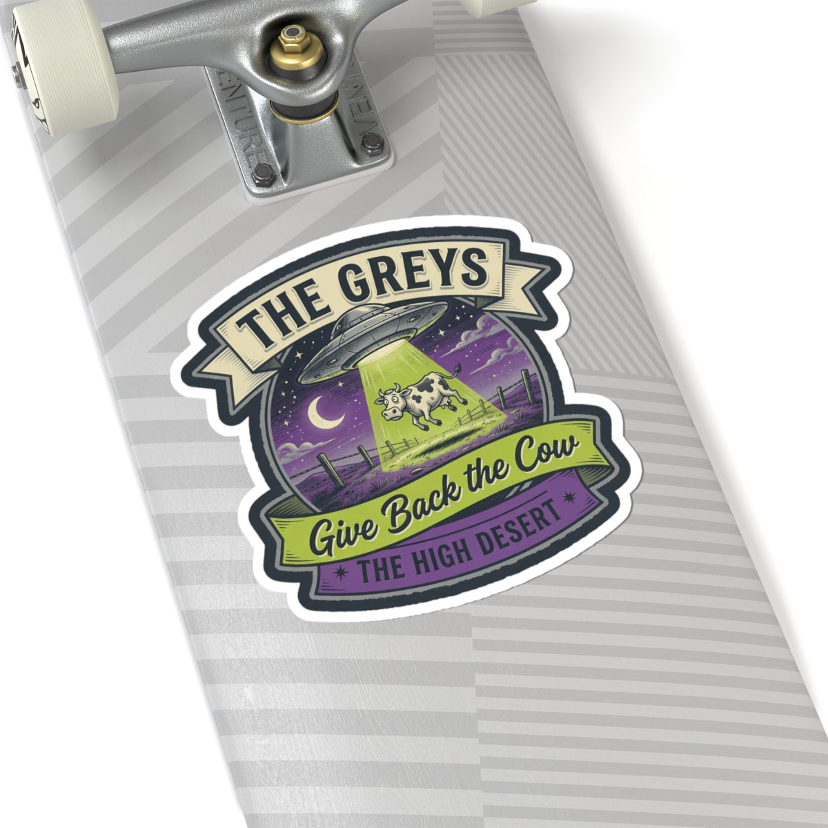 Die-cut sticker on a striped notebook page: UFO and cow, reads 'THE GREYS' and 'THE HIGH DESERT'.