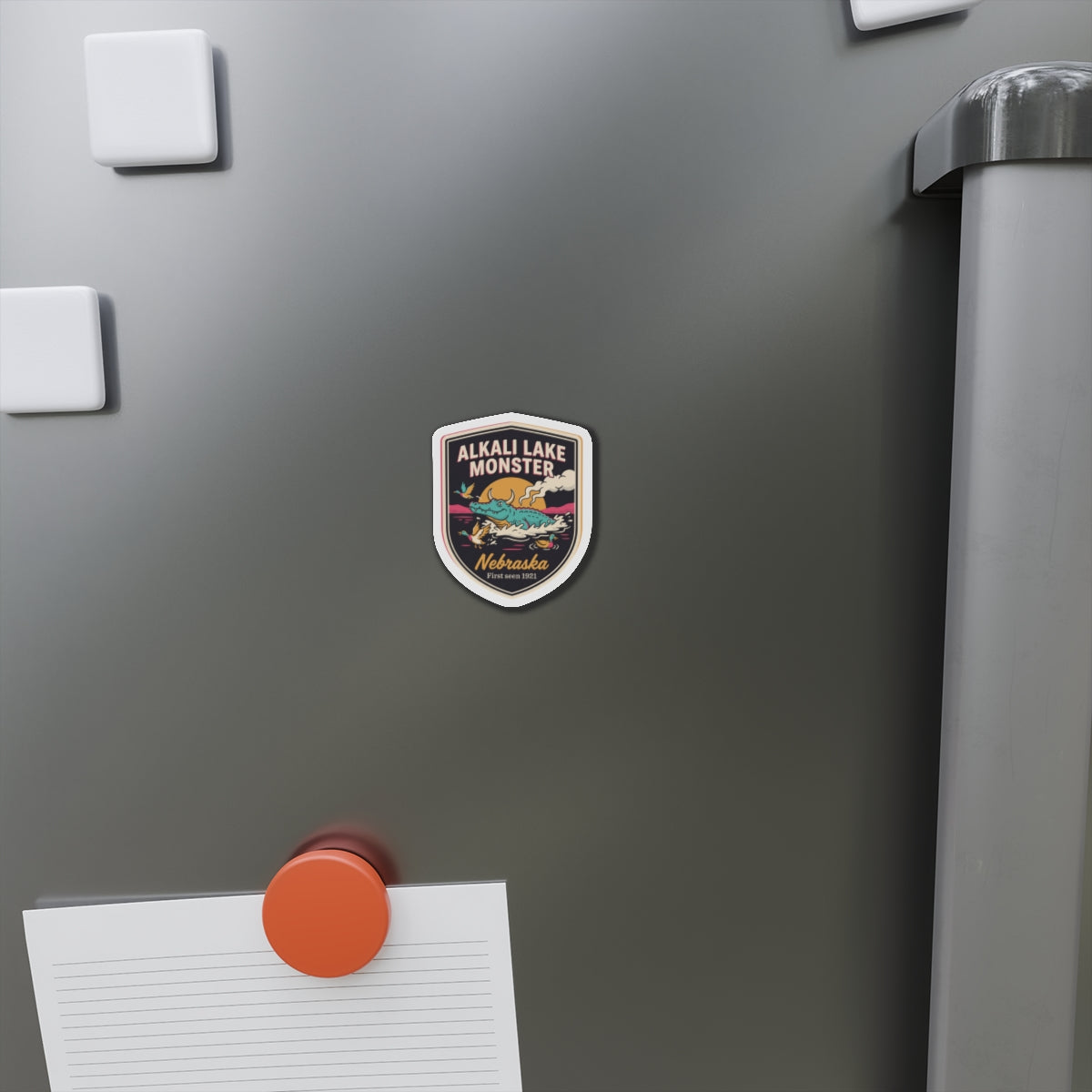 Shield-shaped die-cut magnet on a refrigerator door, featuring Alkali Lake Monster and Nebraska