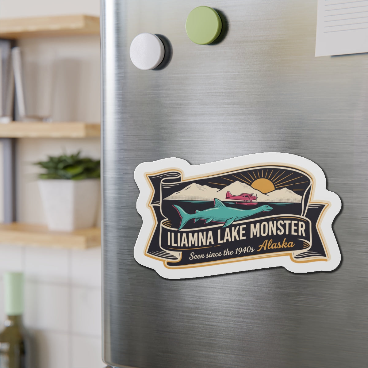 Die-cut magnet sticker with pink and teal lake monster illustration on silver fridge
