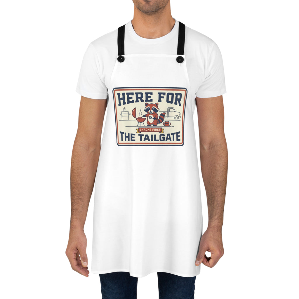 Man wearing white BBQ apron with black straps and 'Here for the Tailgate' raccoon graphic