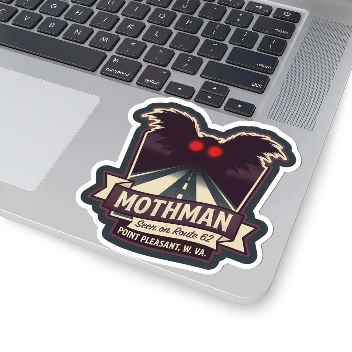 Kiss-cut Mothman sticker on laptop keyboard area, with red eyes and Route 62 graphic