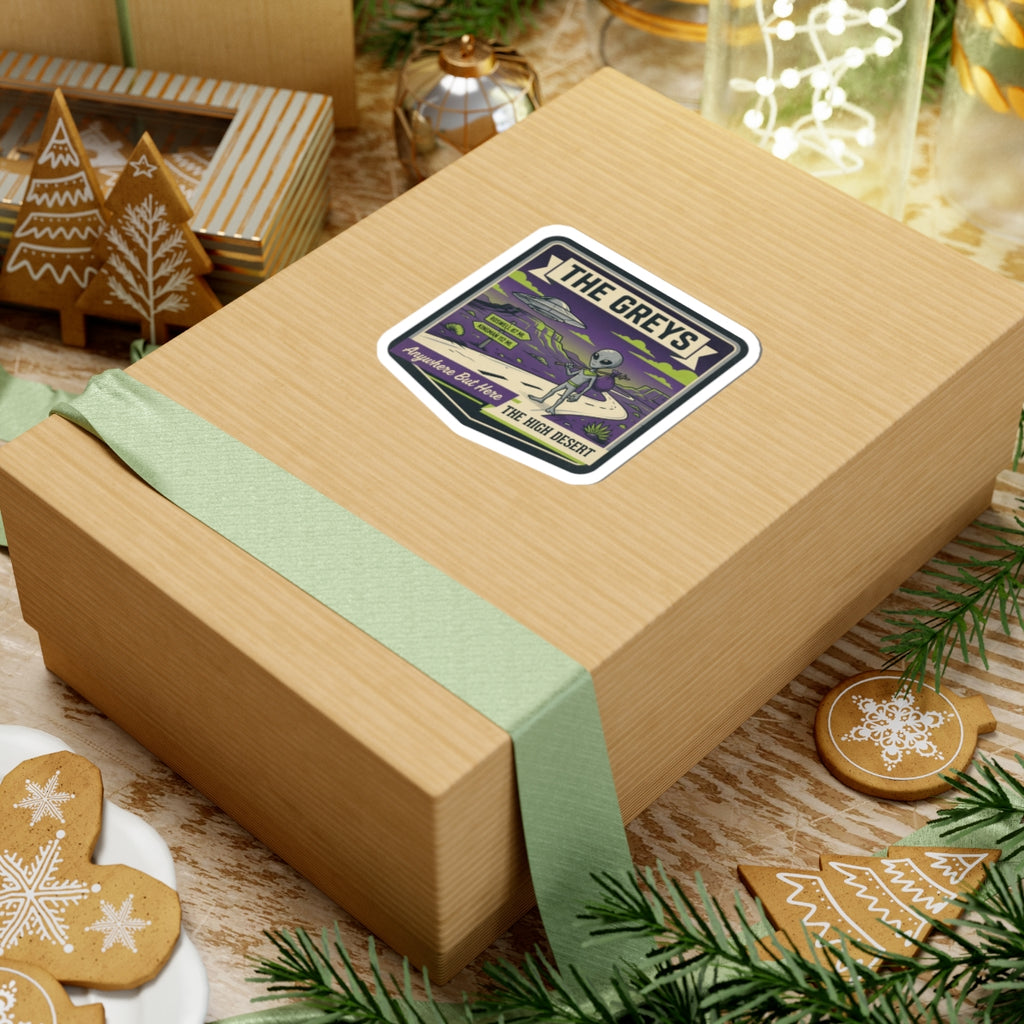 Die-cut sticker on gift box lid with gray alien, UFO, and desert road artwork.
