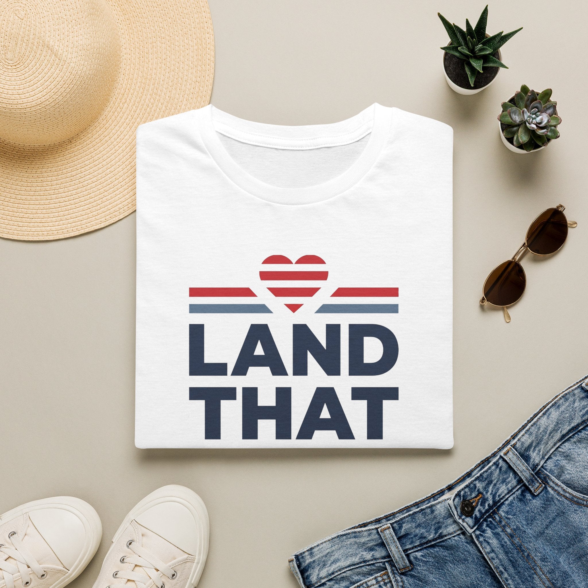 White T-shirt with “LAND THAT” graphic and red heart above blue stripes, crew neck.