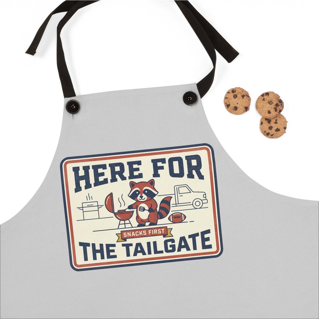 Light gray apron close-up: 'HERE FOR THE TAILGATE' print with raccoon BBQ cartoon