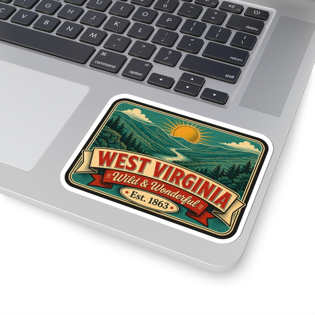 White-bordered vinyl decal on laptop featuring mountains, sun, ribbon “Wild & Wonderful”.