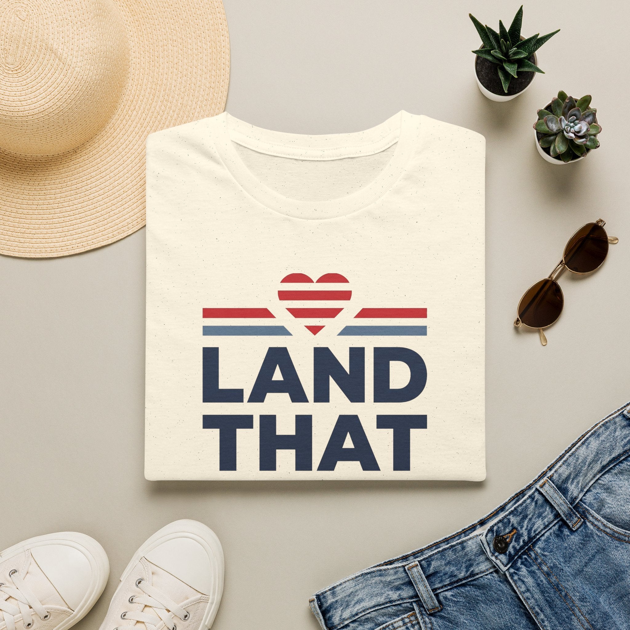 Cream T-shirt with “LAND THAT” graphic and red heart above blue stripes, crew neck.