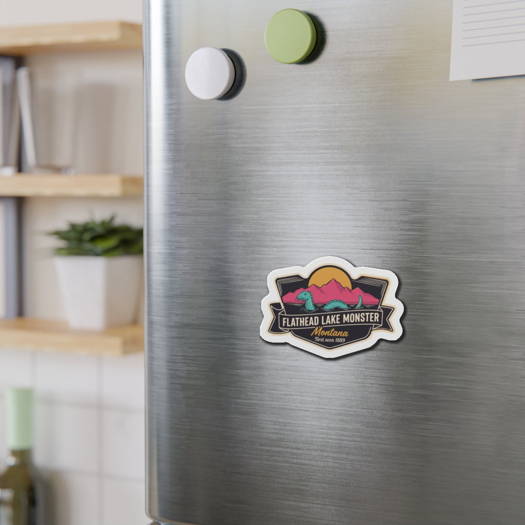 Die-cut Flathead Lake Monster magnet on stainless fridge, teal creature and mountains graphic