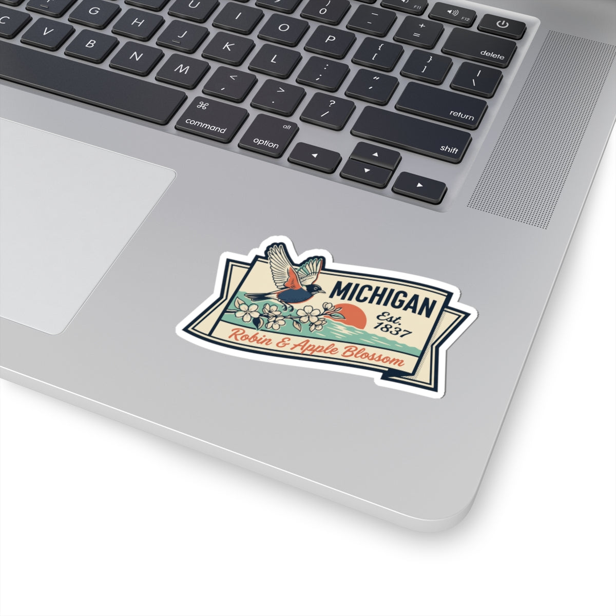 Michigan state badge sticker on laptop touchpad area, showing robin, apple blossoms, “Est. 1837”