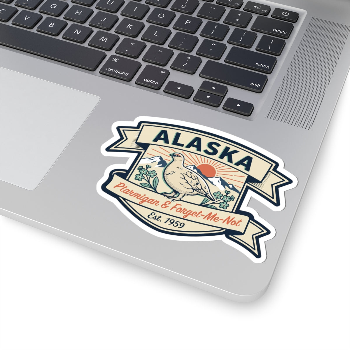 Die-cut Alaska badge sticker on a gray laptop lid with ptarmigan bird and ribbon