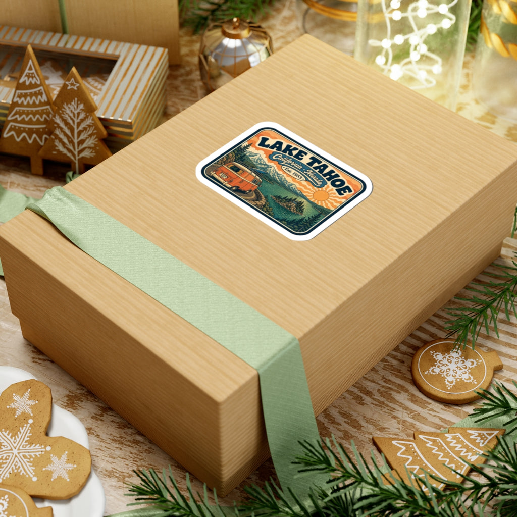 Kiss-cut Lake Tahoe sticker on a wrapped gift box, orange camper van and mountain lake artwork.