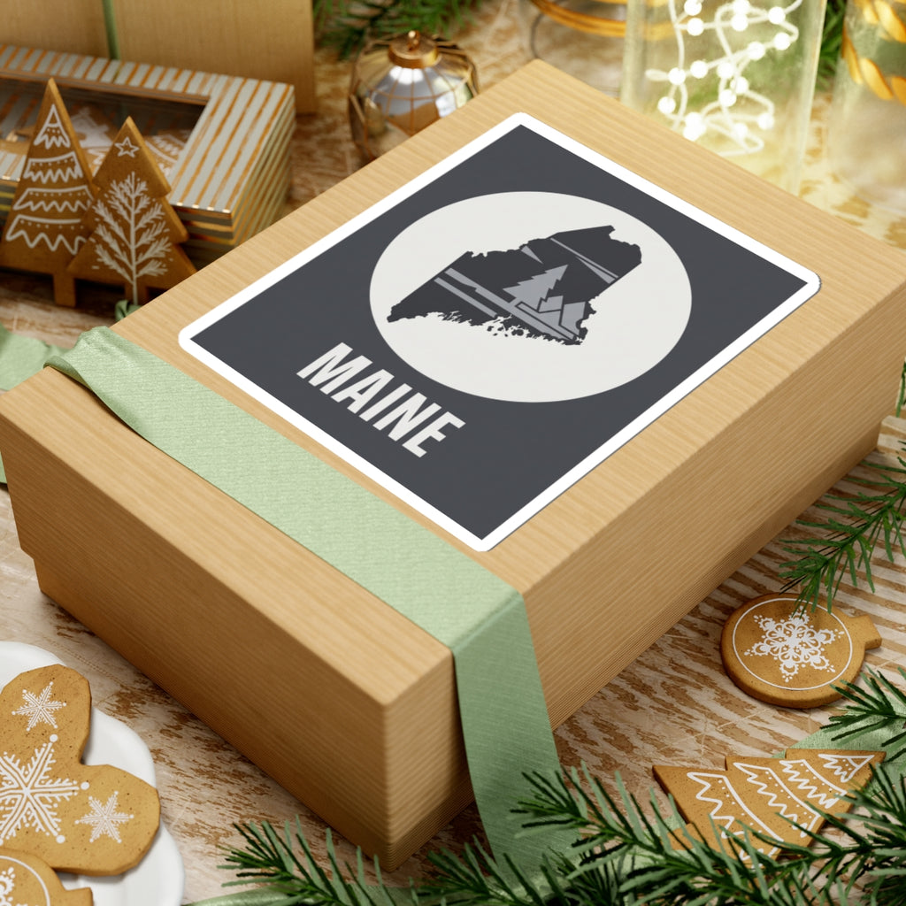 Maine silhouette sticker on gift wrap: dark rectangle, circular pine tree map emblem, 'MAINE'.