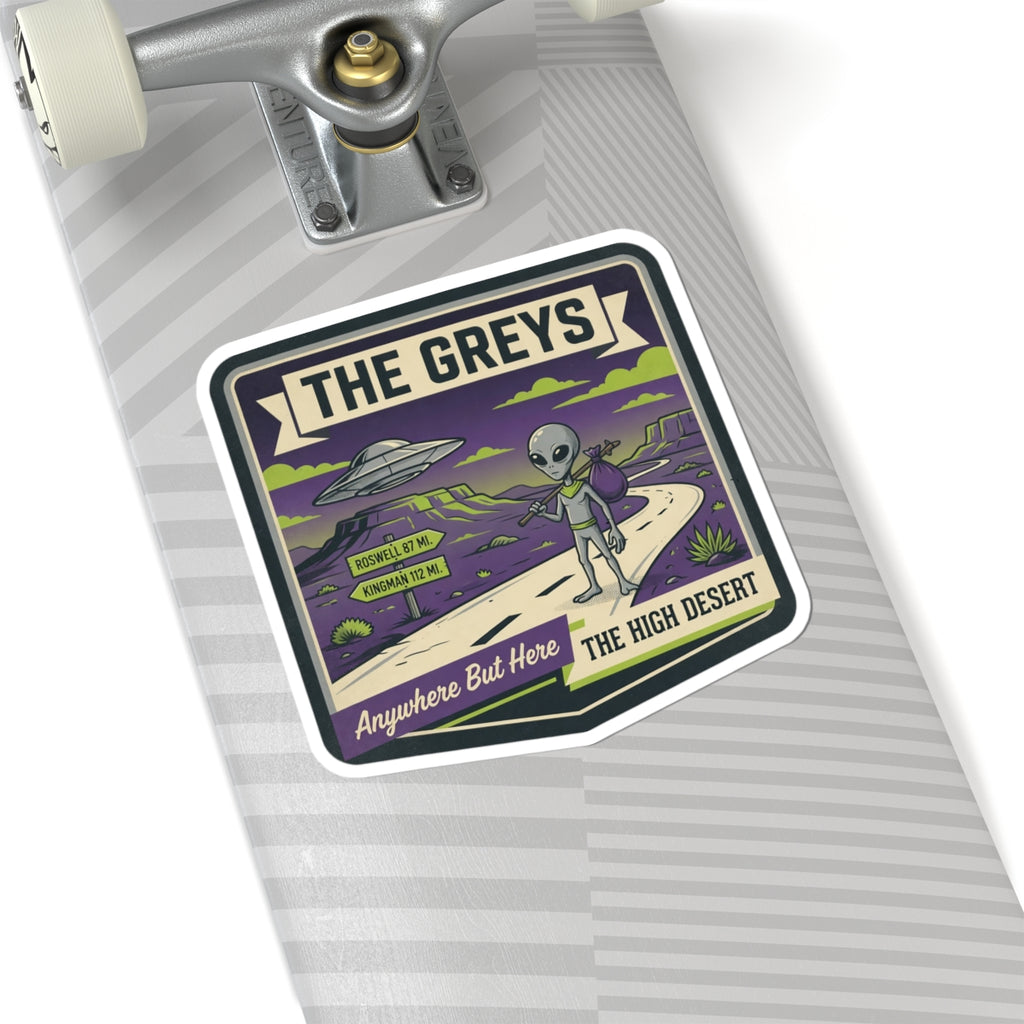 Die-cut sticker on skateboard deck with "THE GREYS" alien, UFO, and desert road.