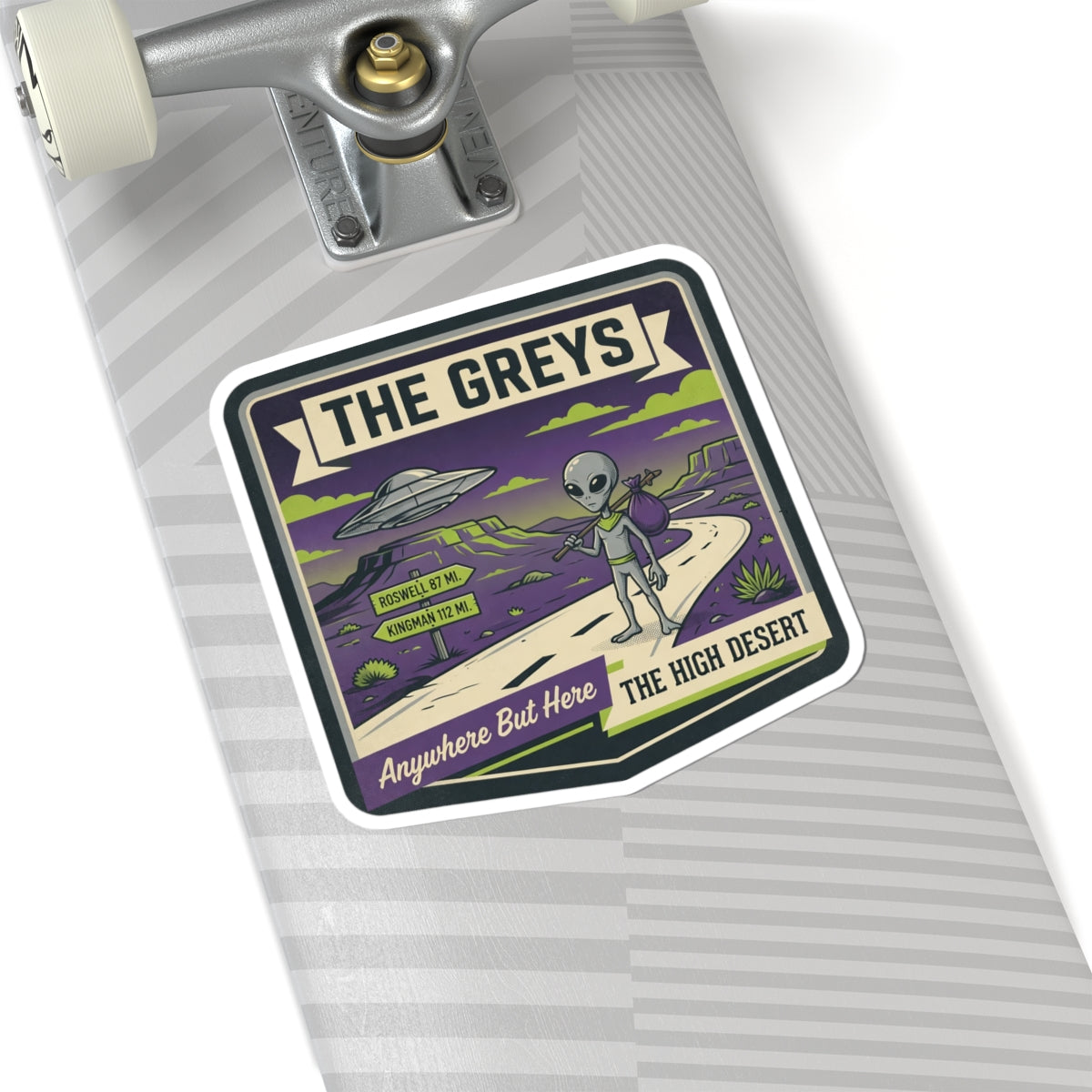 Die-cut sticker on skateboard deck with "THE GREYS" alien, UFO, and desert road.