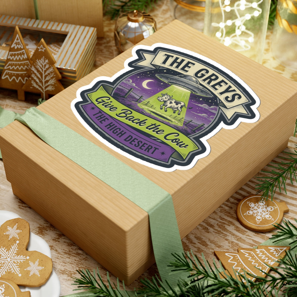 Die-cut sticker on a wrapped gift box with teal ribbon, featuring 'THE GREYS' and 'Give Back the Cow'.
