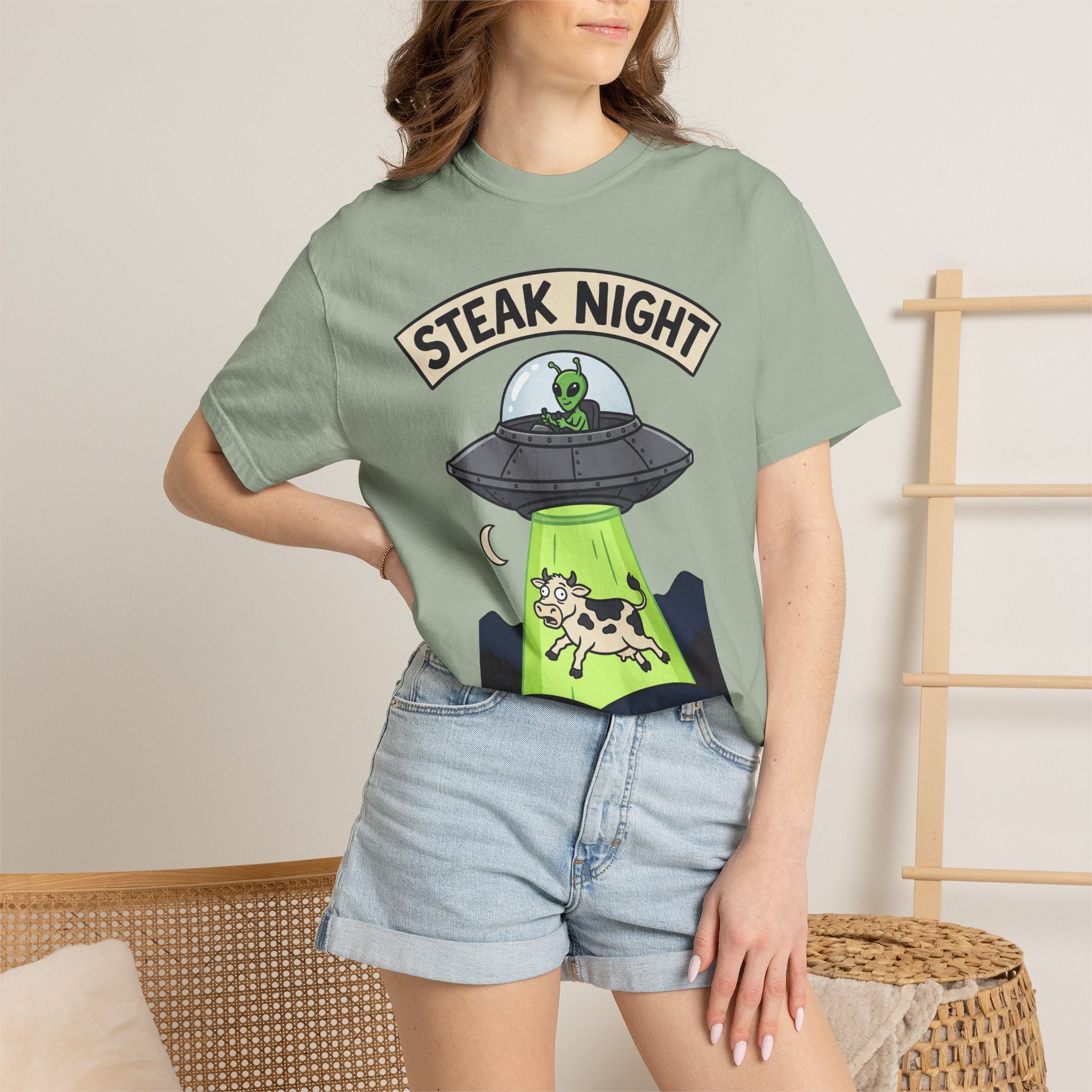 Sage green graphic T-shirt worn by model, 'STEAK NIGHT' UFO and alien cow print.