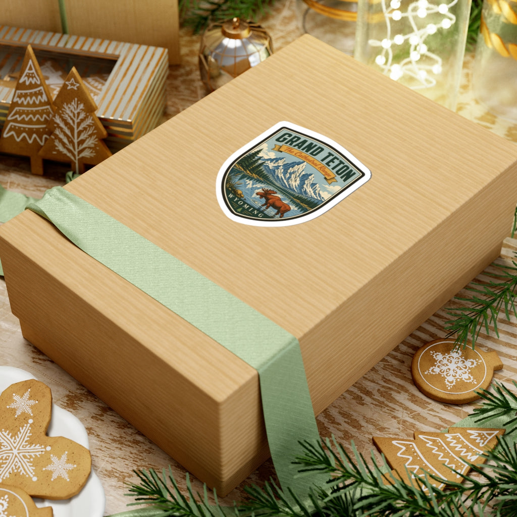 Shield-shaped travel sticker on a wrapped gift box, featuring moose, mountain peaks, and Wyoming text.