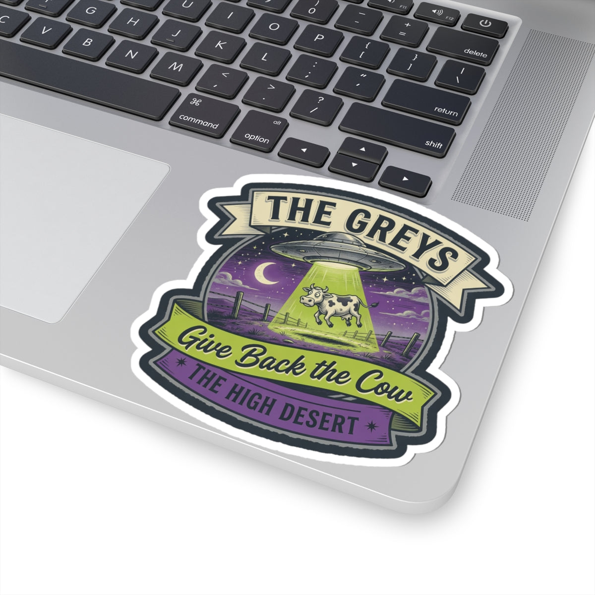 Laptop close-up with die-cut sticker: UFO over cow, 'Give Back the Cow' and 'THE HIGH DESERT'.
