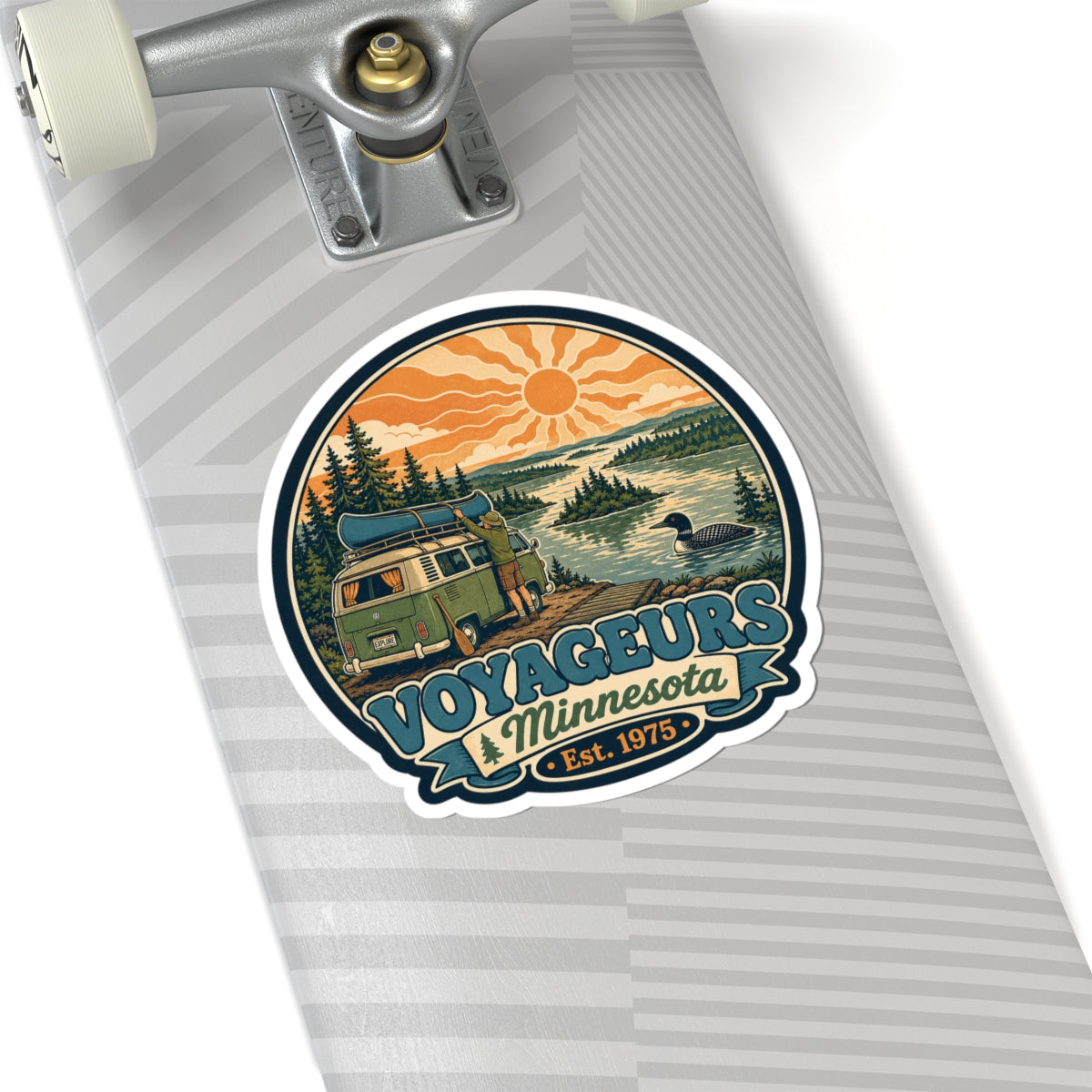 Round die-cut sticker showing green camper van, pine trees, lake and orange sun; Voyageurs Minnesota