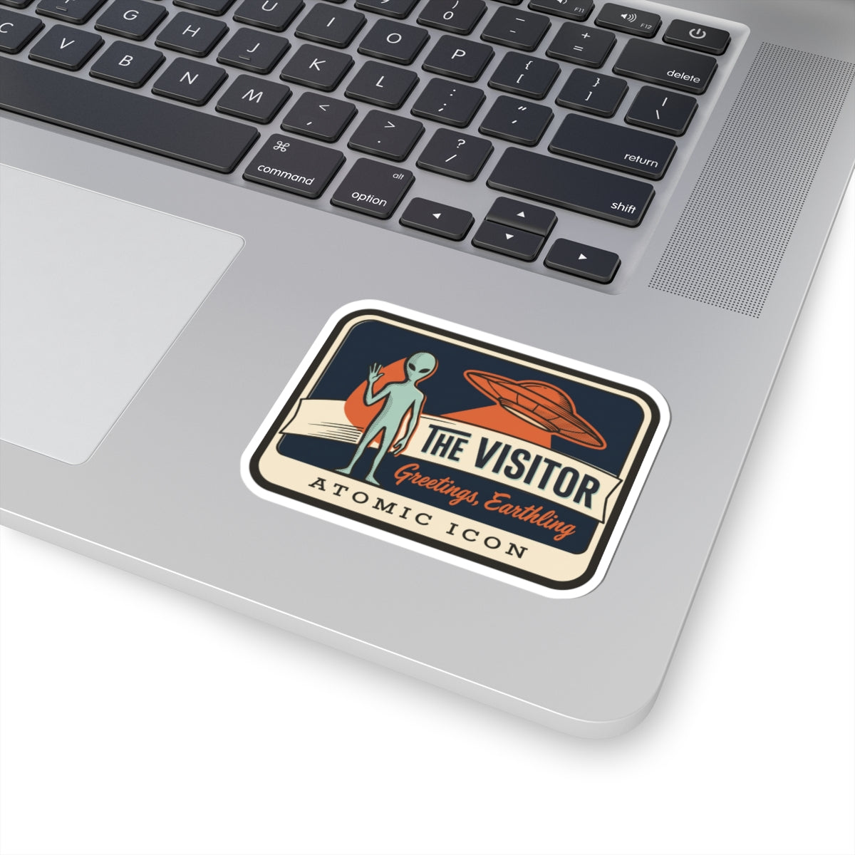 Alien and UFO “THE VISITOR” sticker on a laptop keyboard, “Greetings, Earthling” text