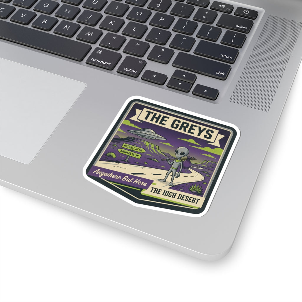 Die-cut sticker on laptop showing "THE GREYS" alien roadtrip desert design.
