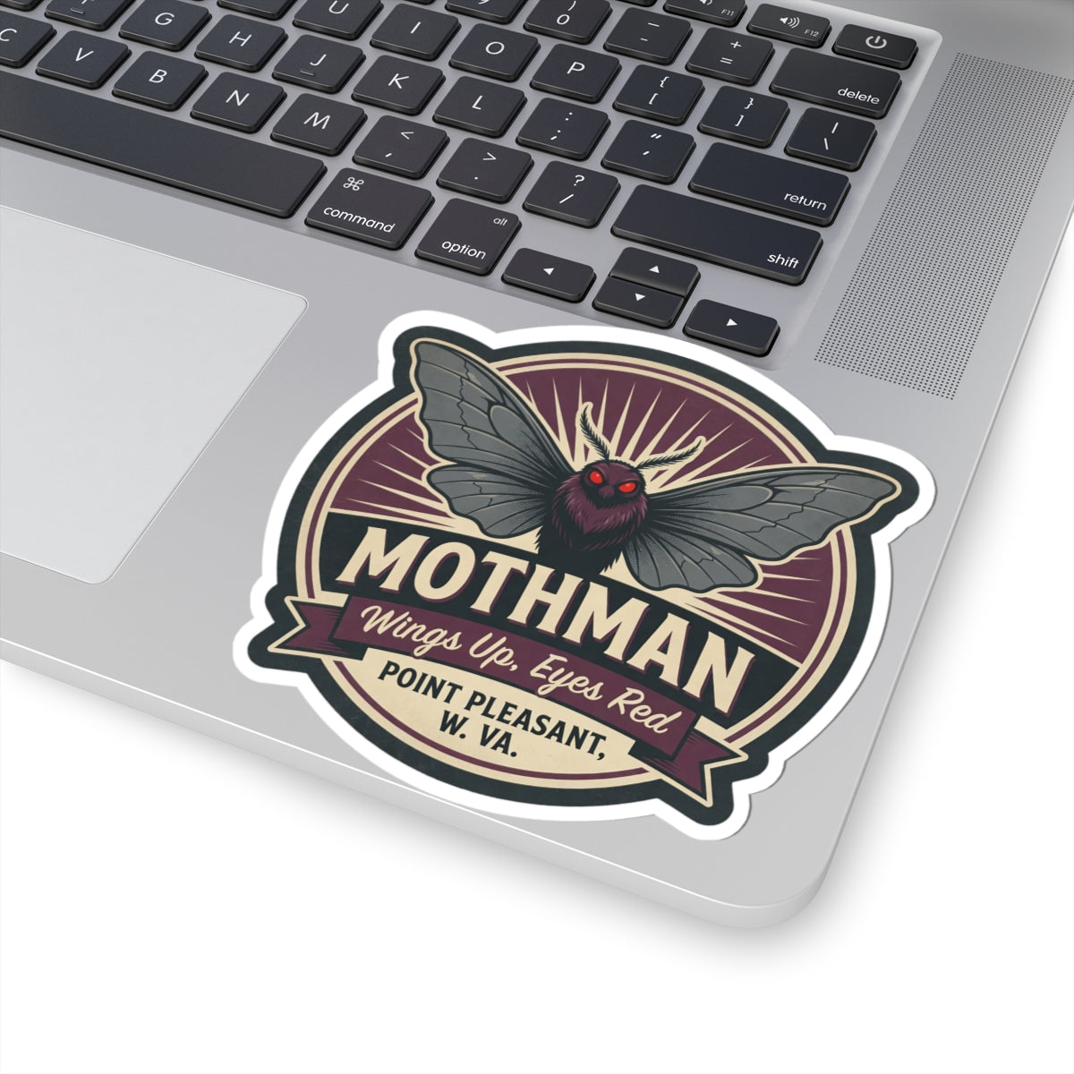 Mothman emblem kiss-cut sticker on laptop surface, close view of wings and red eyes badge