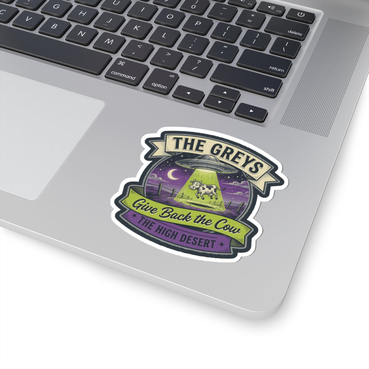Die-cut sticker on a laptop lid: 'THE GREYS' UFO and cow with 'Give Back the Cow' text.